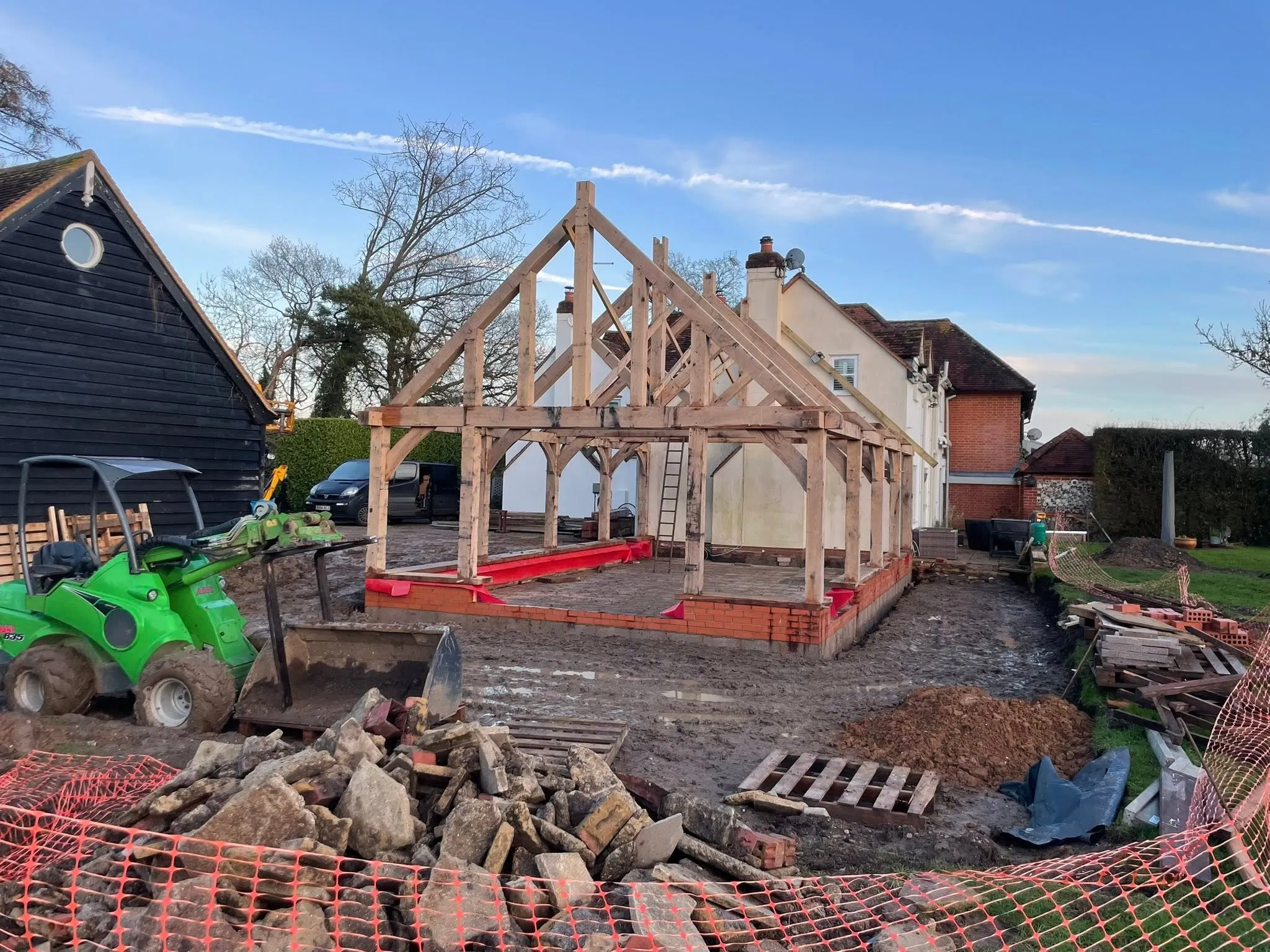 Oak frame raised on its new brick plinth beside the house, with the loader clearing the site