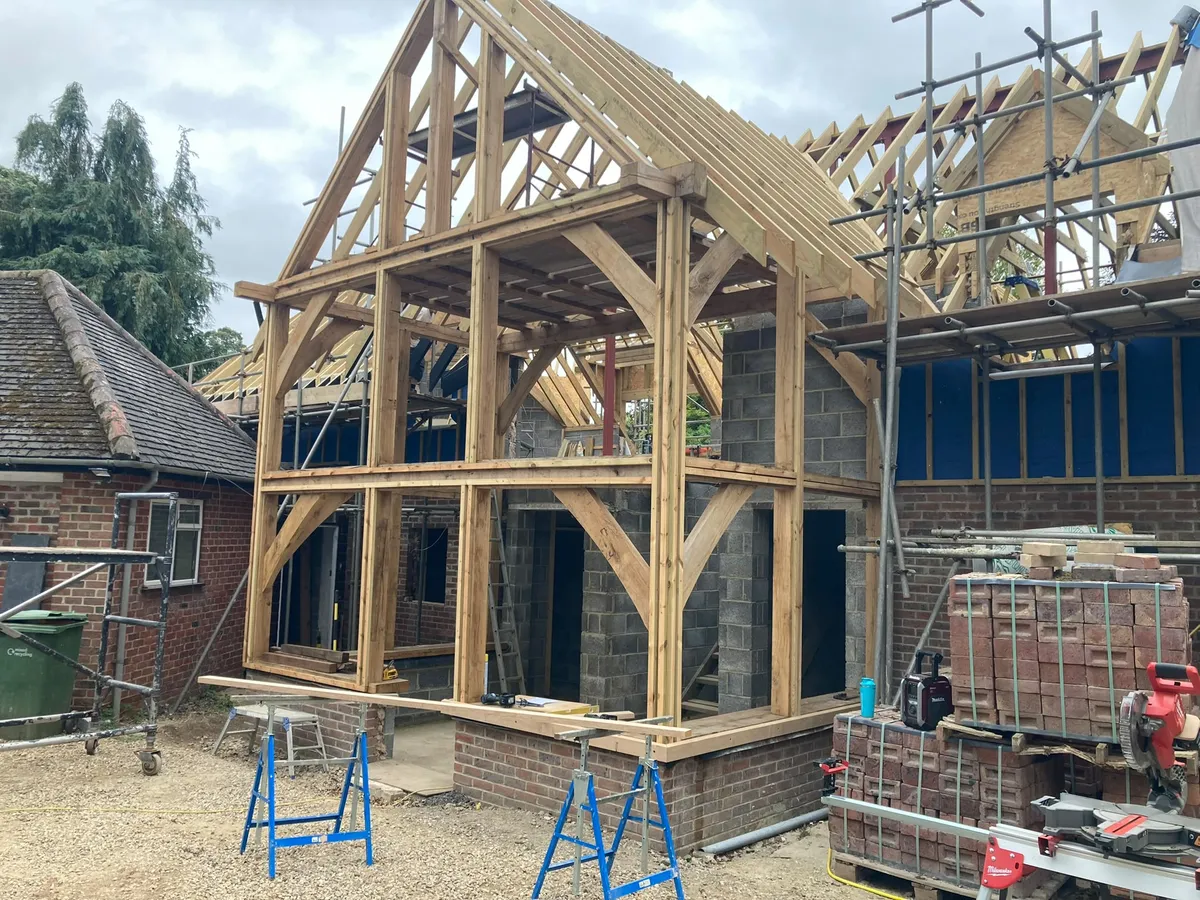 Two-storey oak frame raised on a brick plinth with the rafters in place and scaffolding behind